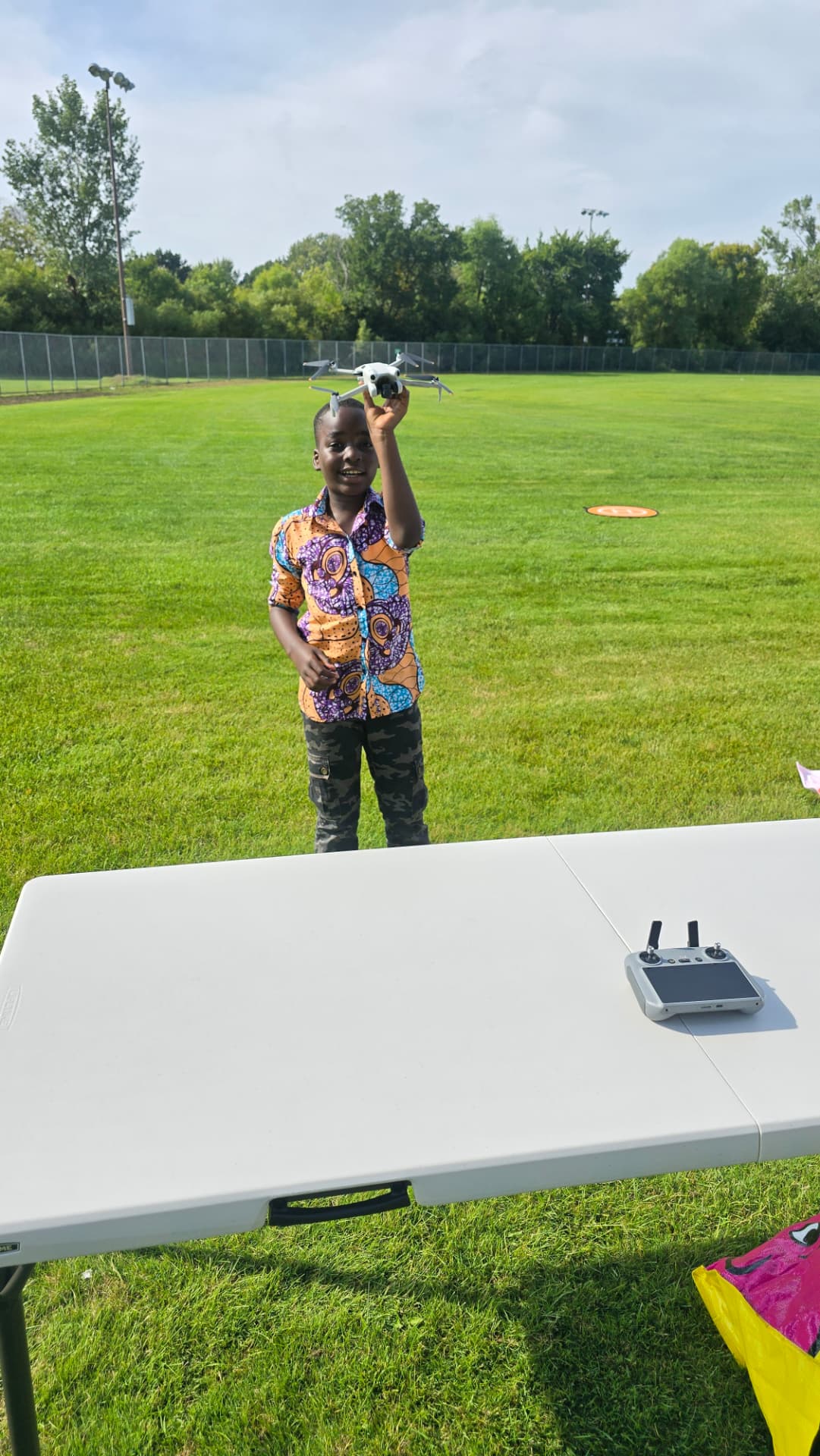 A young participant smiles while holding a small camera drone overhead on a grass field, with a controller resting on a table nearby.